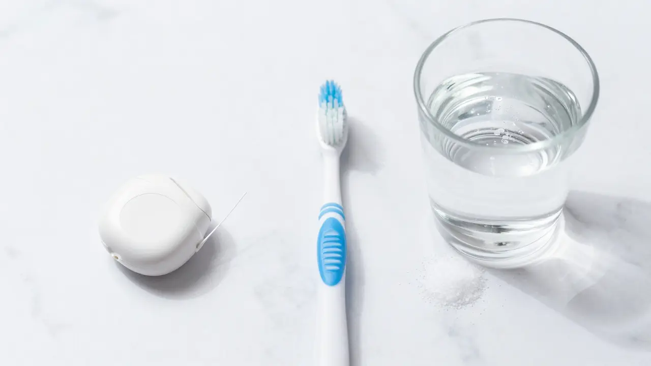 Měkký zubní kartáček, nit a sklenice slané vody na mramorovém podkladu.